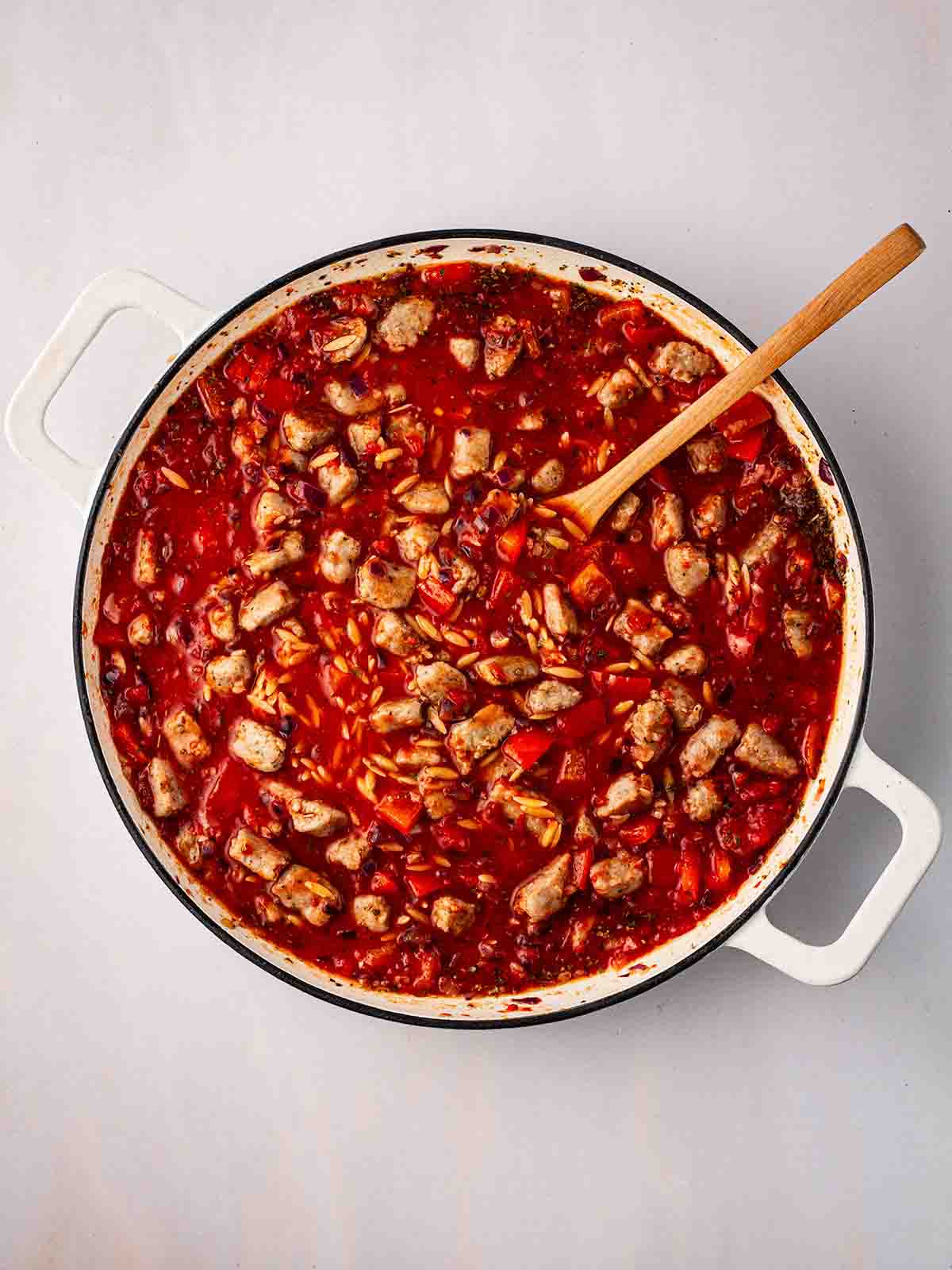 Chunks of sausage and red pepper in a pan with orzo and tomato sauce, with a wooden spoon for for step 3 in the recipe for Sausage Orzo.