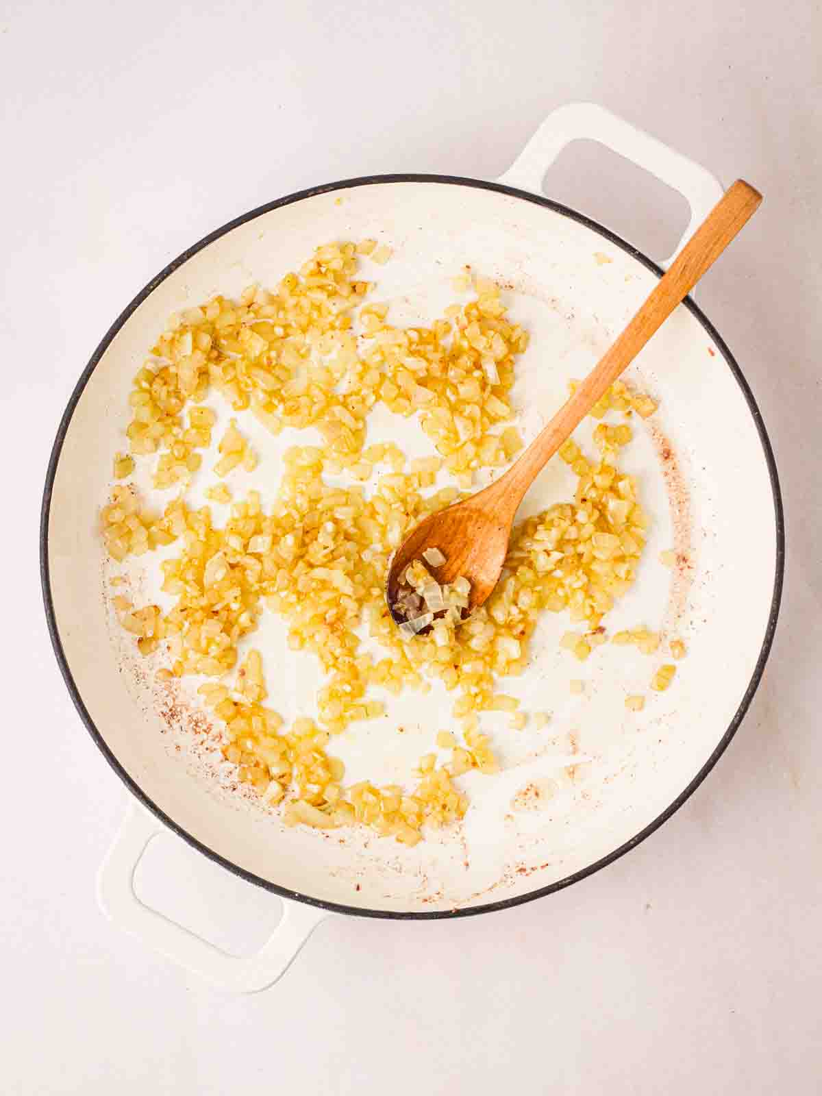Diced onions frying in a white pan with a wooden spoon to stir for step 3 in the recipe for Spaghetti Bolognese.