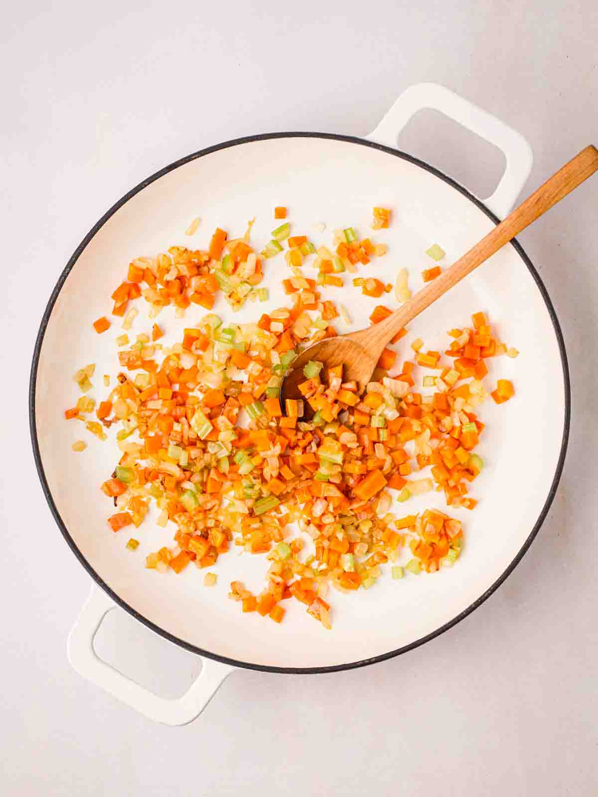 Diced onions, carrots and celery in a white pan, being stirred with a wooden spoon, for step 4 in the recipe for Spaghetti Bolognese.