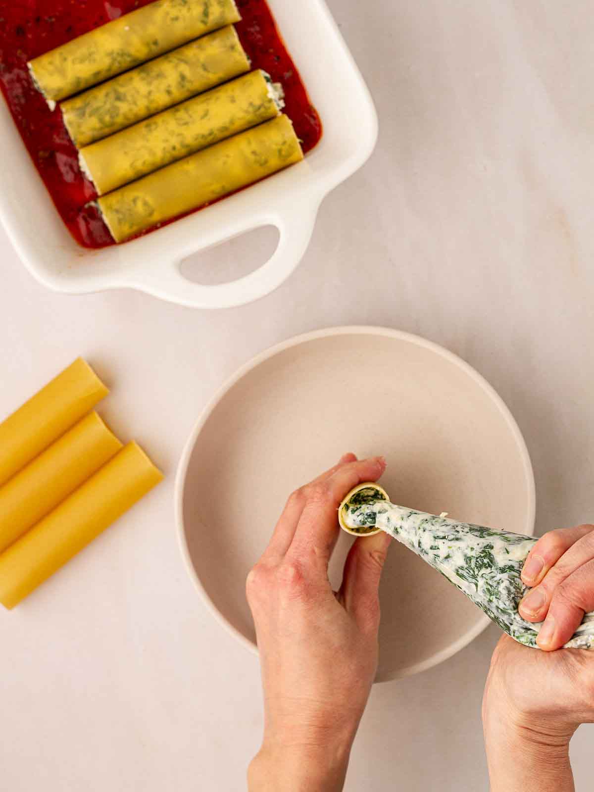 Two hands filling cannelloni tubes with spinach and ricotta filling, with a dish of already filled tubes.
