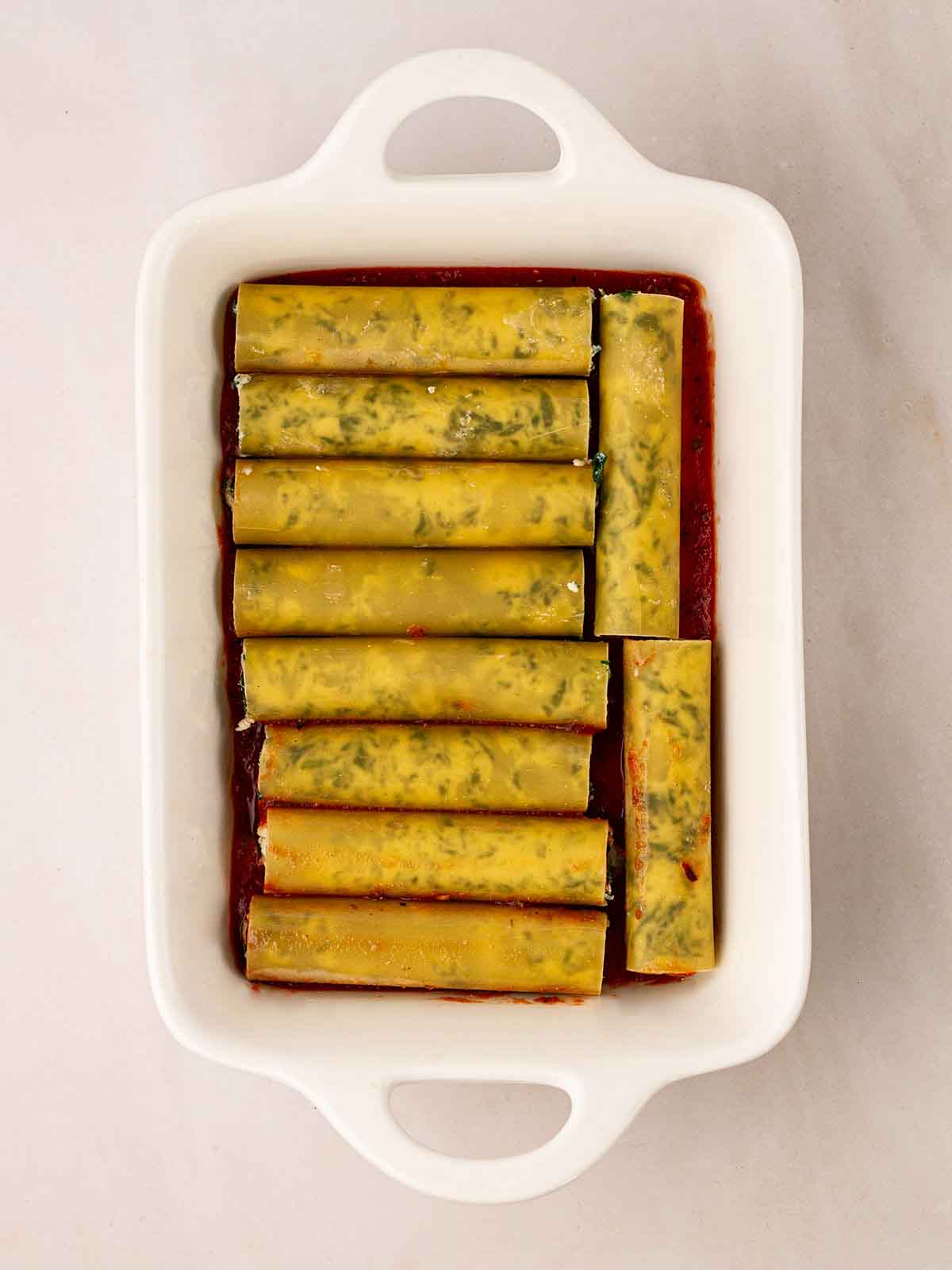 Unbaked cannelloni tubes filled with spinach and ricotta on a bed of tomato sauce for step 6 in the recipe for Spinach and Ricotta Cannelloni.