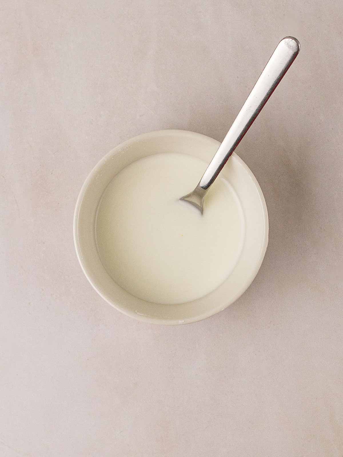 A small white bowl of white liquid - cornflour and water - and a spoon stirring, to thicken Sweet and Sour Chicken sauce.