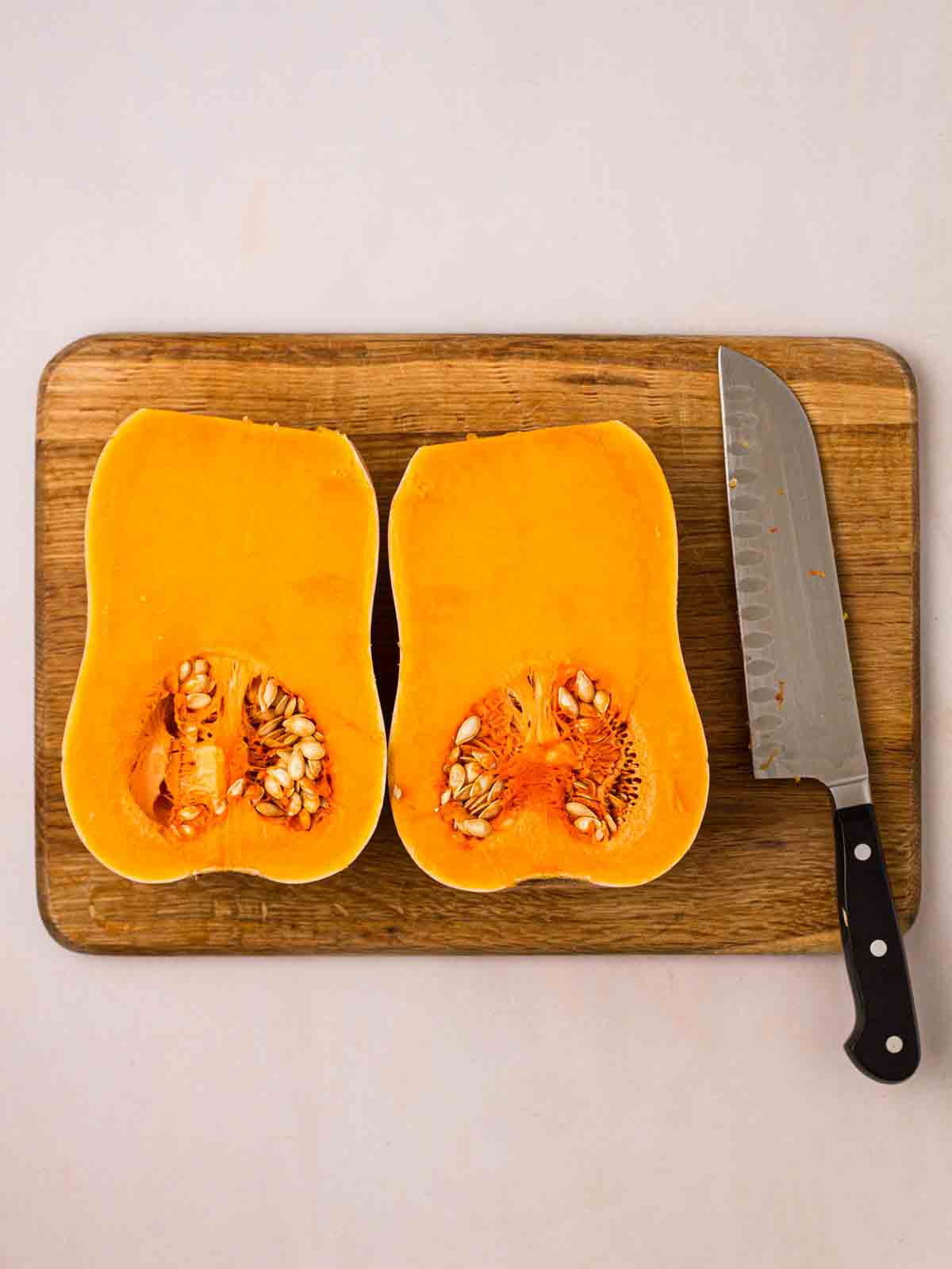 A halved butternut squash on a chopping board with a knife to the side.