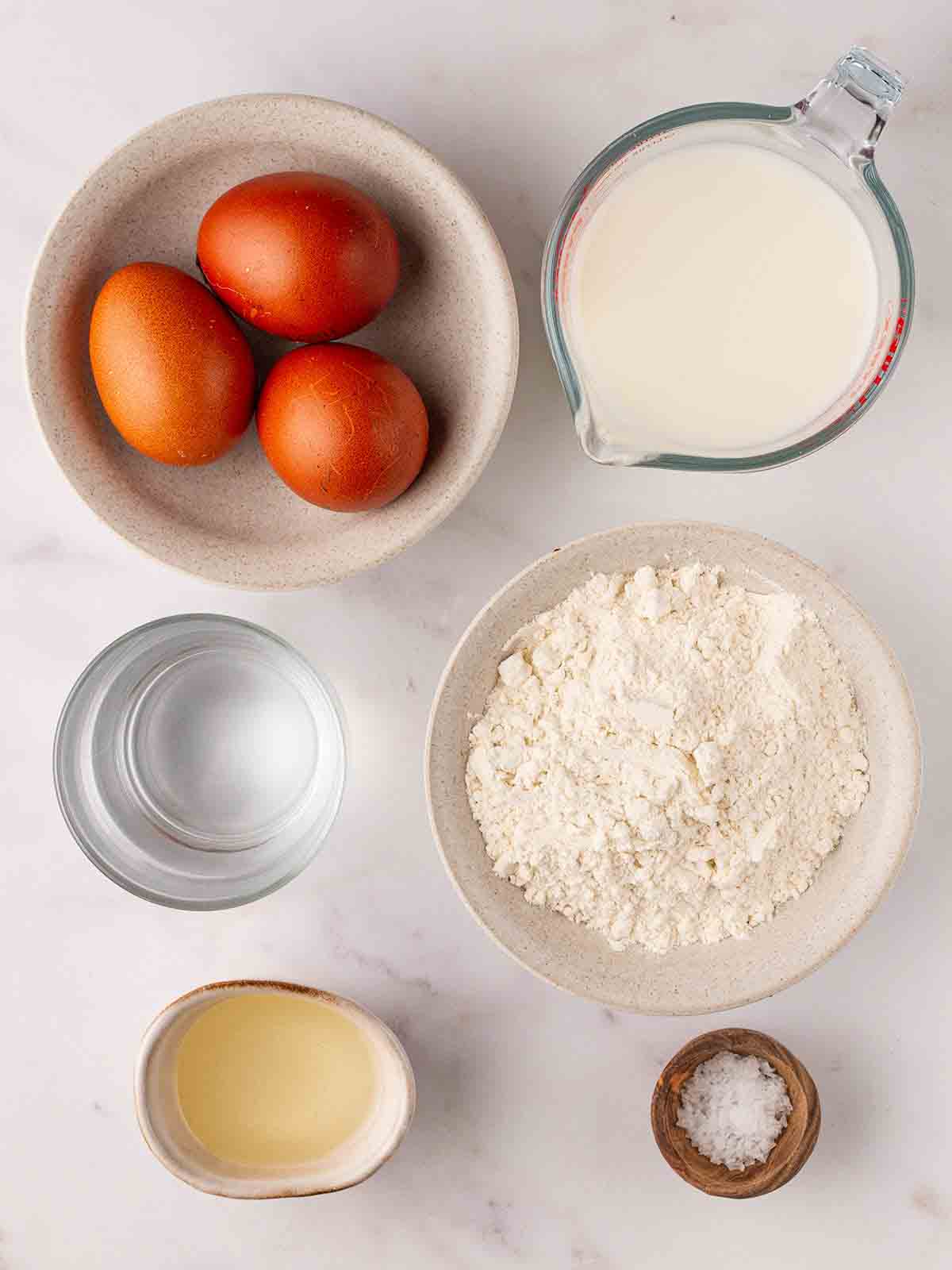 The ingredients for Yorkshire Puddings laid out on a counter top. Three eggs, milk, plain flour, salt, oil and water.
